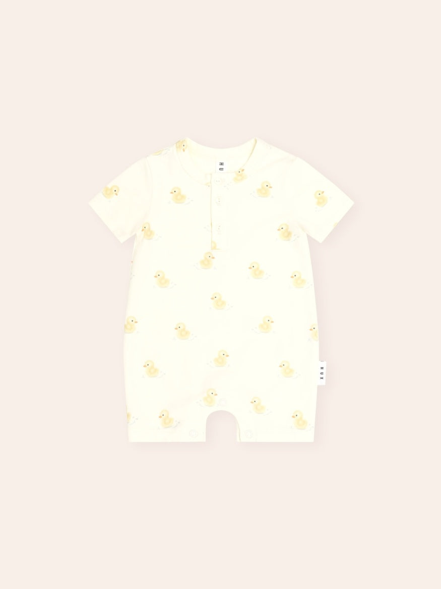 Cream short-sleeve baby Henley bodysuit with an all-over yellow rubber duck print. Features a three-button Henley neckline, soft cotton fabric, and snap closures at the crotch for easy nappy changes. Lightweight and perfect for everyday wear in warmer weather.