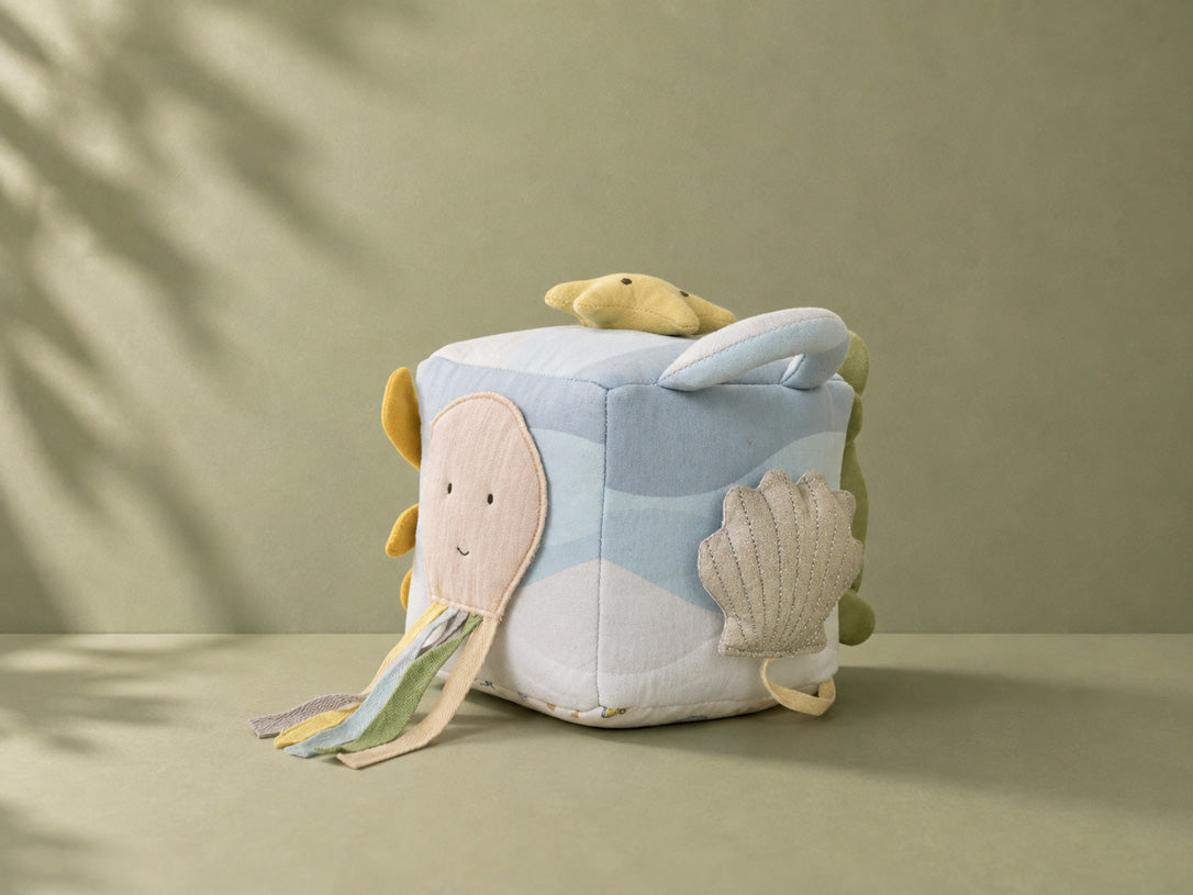 A cube-shaped plush activity cube toy with an ocean theme, featuring a light blue and white wavy pattern, a sewn-on smiling octopus, a shell applique, and a yellow star on top, set against a neutral olive-green background with soft leaf shadows.