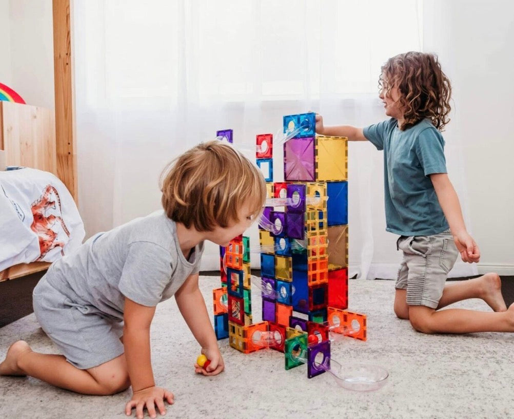 Two children building a tall tower from colourful magnetic tiles on a living-room floor